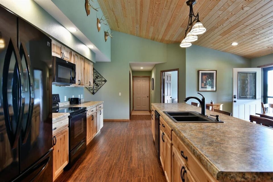 Modern kitchen with light wood cabinets, a black refrigerator and stove, a large center island with a double sink, and light green walls under a wood-plank ceiling.