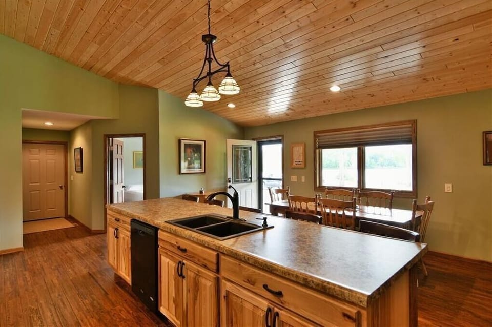 Modern kitchen with light wood cabinets, a black refrigerator and stove, a large center island with a double sink, and light green walls under a wood-plank ceiling.