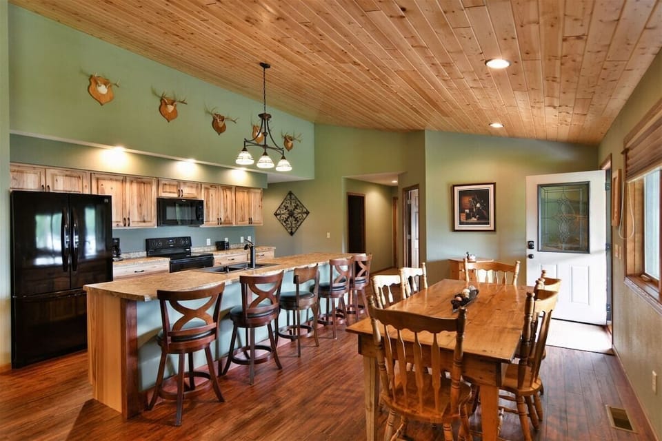 An open-concept kitchen and dining area featuring a large wooden island with bar stools, a matching dining table, and rustic wood-paneled ceilings with recessed lighting.