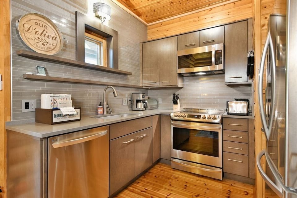 A modern, compact kitchen in The Bike Cabin featuring light wood cabinetry, stainless steel appliances, a gray tiled backsplash, and wooden ceilings.