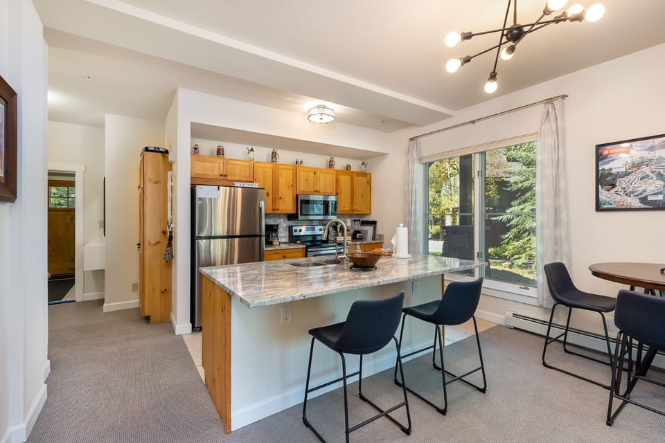 Modern kitchen with stainless steel appliances, wooden cabinets, and a marble island countertop with four black barstools. There is a small round dining table in the corner and large windows.