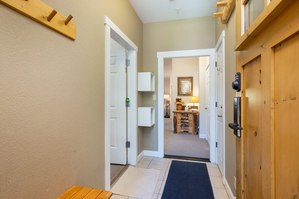 A hallway view featuring a wooden coat rack and white door on the left, leading to a room with a lamp and wooden furniture. A wooden entrance door is slightly open on the right.