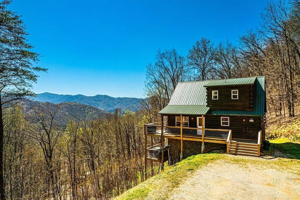 Log cabin exterior surrounded by wooded hillsides with long-range mountain views near Bryson City. Gravel driveway provides direct cabin access.