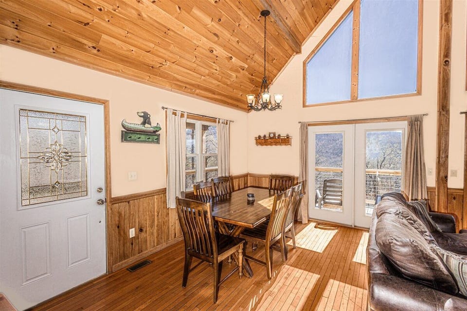 Dining table positioned beside large windows and exterior door within the open main level. Wood ceilings and wall accents complement the cabin setting.