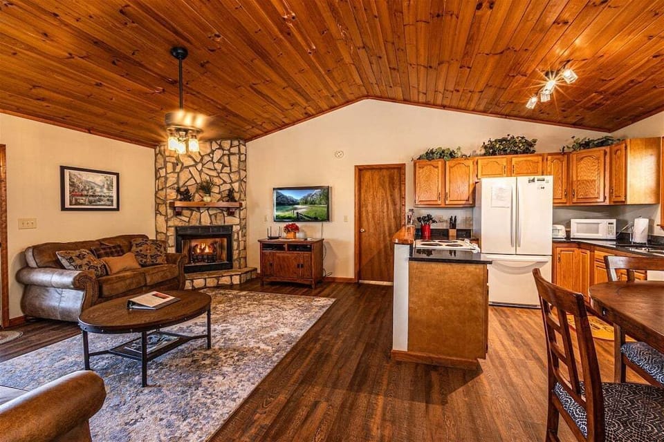 Open-concept living room and kitchen featuring a stone fireplace, wood-paneled ceiling, and dining area. Comfortable indoor gathering space for a Smoky Mountains cabin stay near Bryson City.