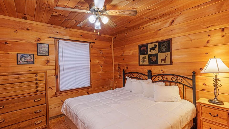 The bedroom includes a wrought iron bed with white bedding, bedside tables, and a ceiling fan. Wood walls create a classic Smoky Mountain cabin feel.
