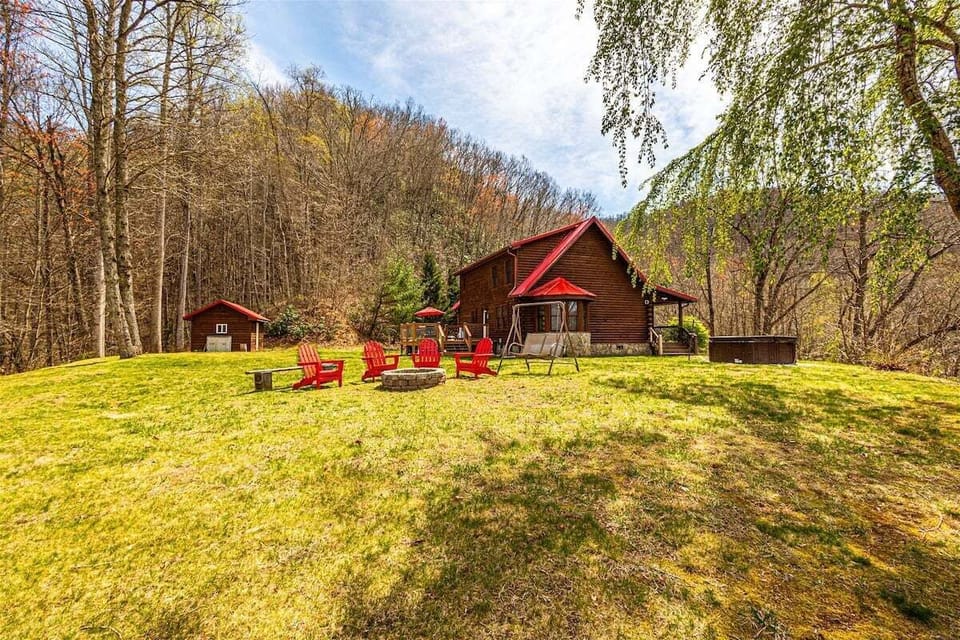 A wide grassy yard stretches between the main cabin, a small outbuilding, and red outdoor chairs. Surrounding woods create privacy at this Smoky Mountains rental.