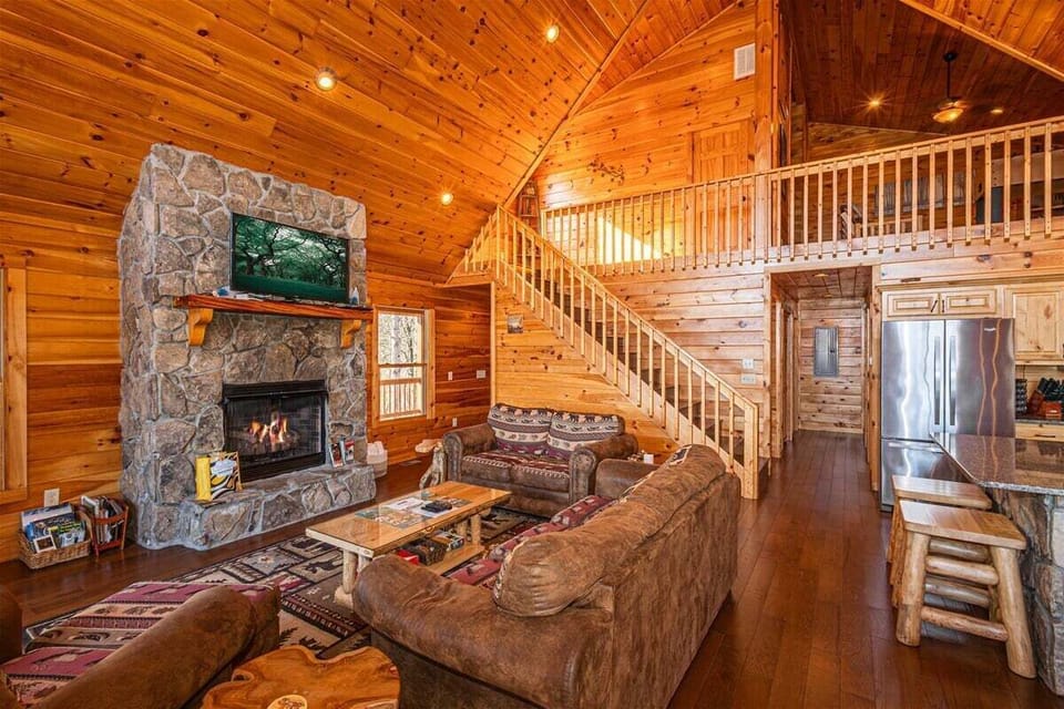 The living room features vaulted wood ceilings, a stone fireplace, loft railing, and open access to the kitchen. Large windows frame mountain views throughout the space.