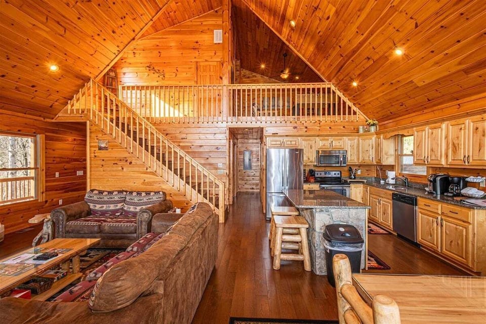 The open floor plan connects the living area, dining space, staircase, and fully equipped kitchen beneath vaulted wood ceilings. The layout offers ample gathering space for guests.