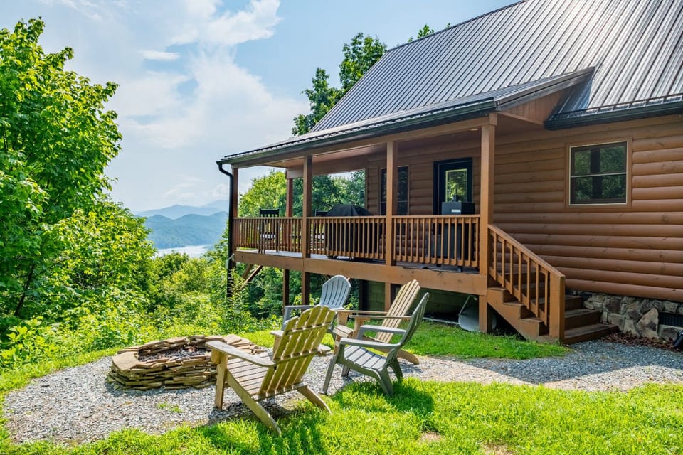 The log cabin exterior features a covered porch, Adirondack chairs, and a stone fire pit surrounded by mountain scenery. Guests can enjoy outdoor gathering space with wooded surroundings.