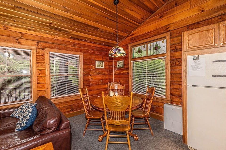 Round dining table positioned beside multiple windows with forest views and adjacent kitchen access. Warm log cabin finishes create a cozy dining space in Bryson City.