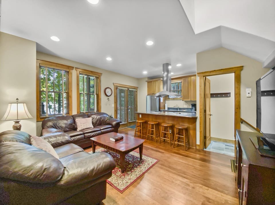 A cozy living room with leather sofas, a coffee table, and a rug, adjoining a kitchen with wooden cabinets, an island with bar stools, and recessed lighting. Large windows let in natural light.