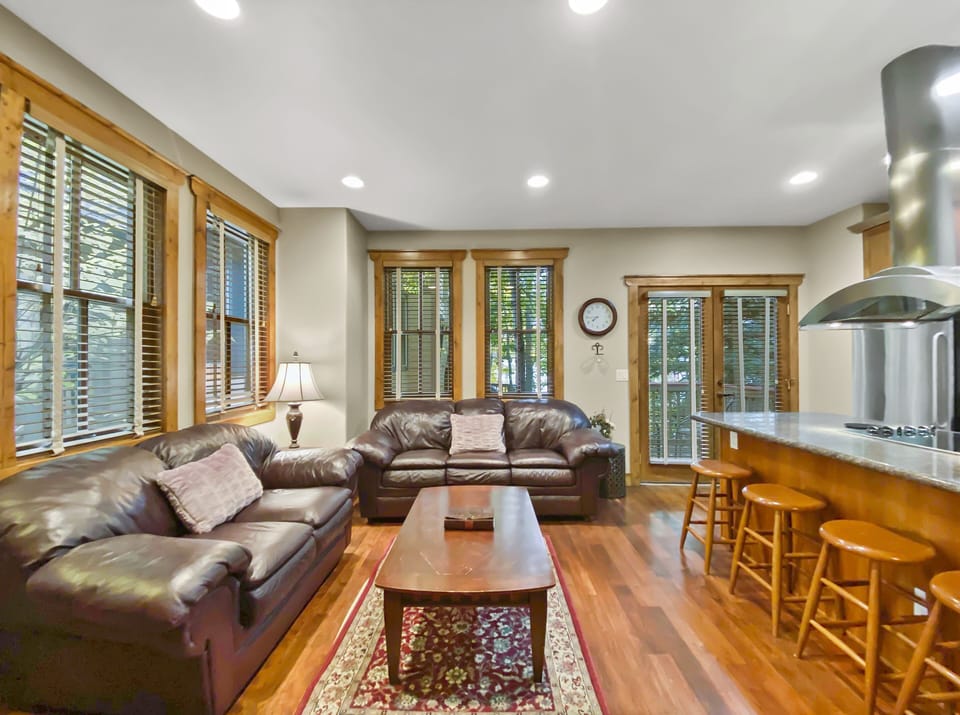 A living room with two brown leather sofas, a wooden coffee table, and a kitchen area with bar stools. The room has wooden flooring and large windows with blinds.