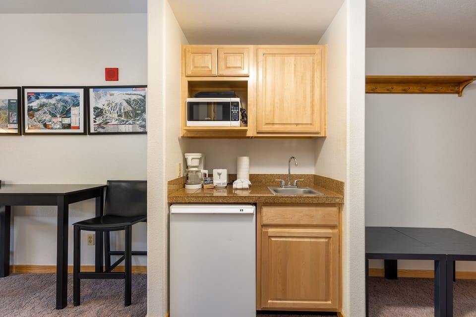 A compact kitchenette with a mini fridge, microwave, sink, coffee maker, and wooden cabinets. Adjacent is a black table and chair, and a coat rack on the right side. Artwork is hung on the left wall.