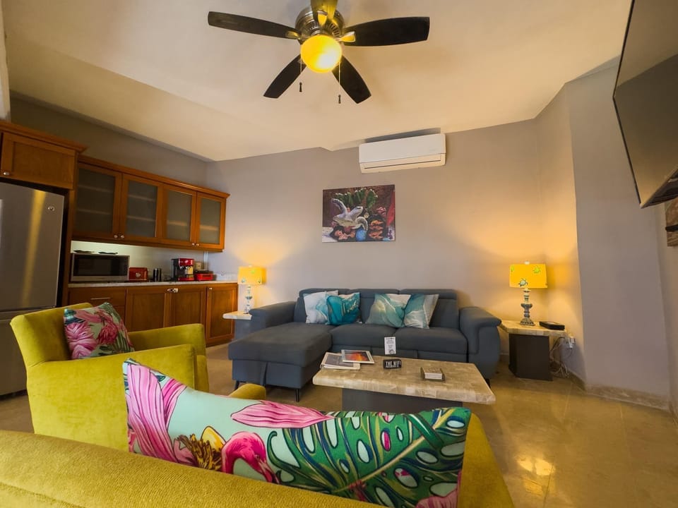 Living area with gray sectional, yellow armchairs accented by floral pillows, marble coffee table, and ceiling fan light.