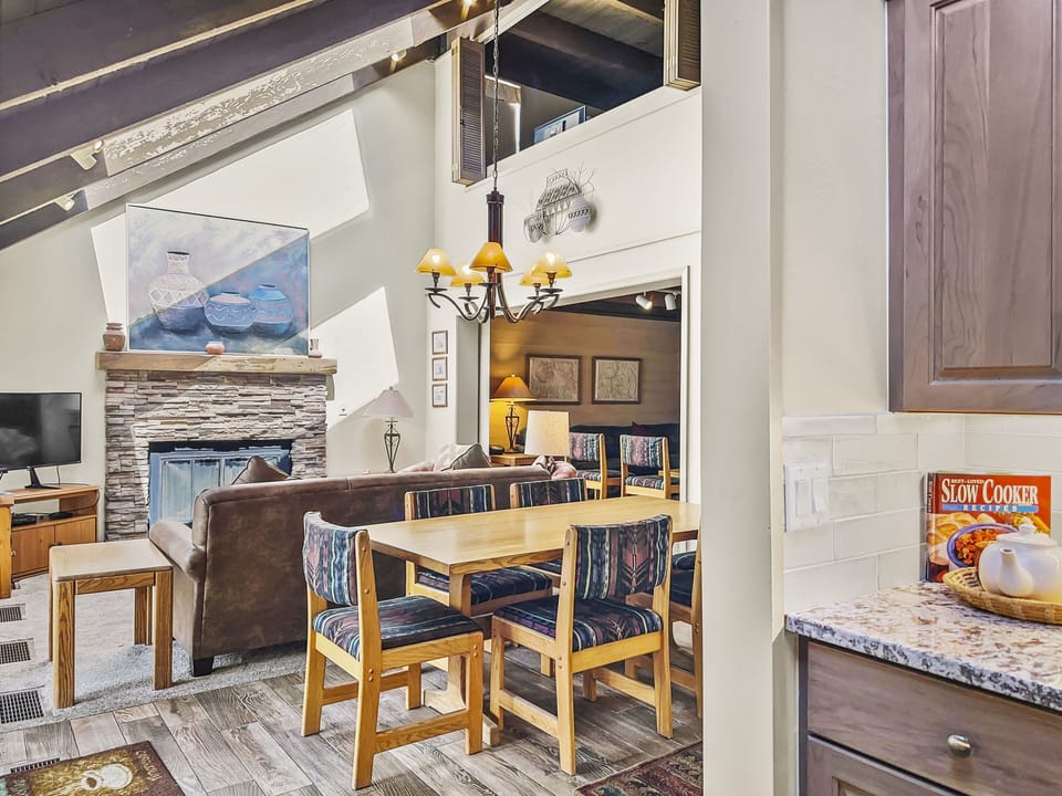 A modern open-concept living and dining area with a fireplace, wooden furniture, wall art, and a hanging chandelier. The kitchen counter is visible in the foreground with a book and a fruit bowl.