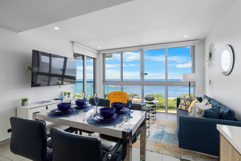 Your sunlit living and dining space frames stunning ocean views through floor-to-ceiling windows. Modern elegance meets coastal comfort with chic furnishings, inviting you to dine, relax, and soak in the serene blue horizon.