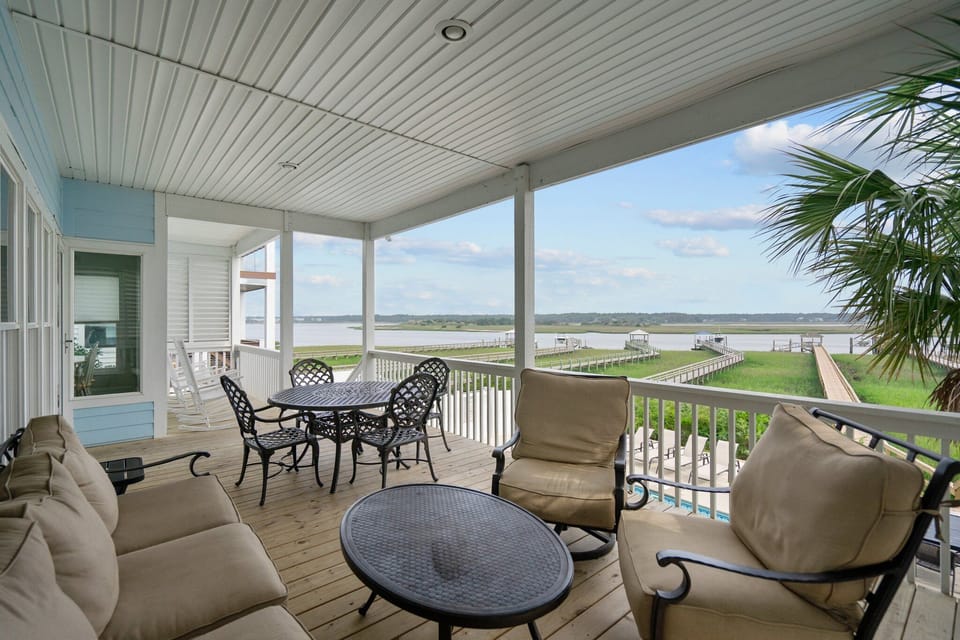Covered porch with cushioned seating, outdoor dining tables, white railings, marsh and waterway views, pool below, palm tree, and wide coastal scenery.