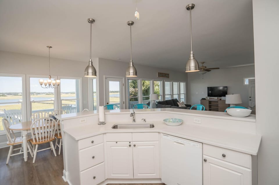 Bright white kitchen with breakfast bar, pendant lights, stainless appliances, ceiling fan, wood-look floors, large windows, and beautiful waterway views.