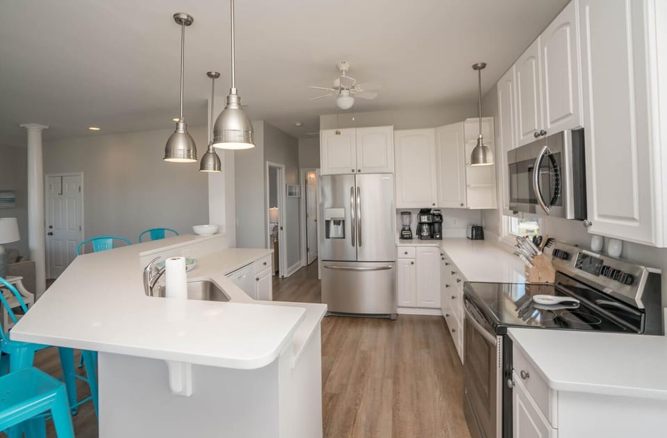 Bright white kitchen with breakfast bar, turquoise stools, stainless appliances, pendant lights, coffee station, ceiling fan, and wood-look floors.