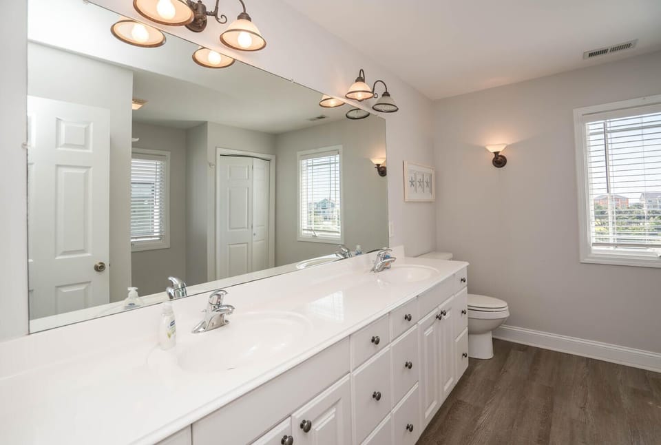 Bright bathroom with long double vanity, large mirror, warm wall lights, toilet, window, gray walls, wood-look floors, and clean coastal style.
