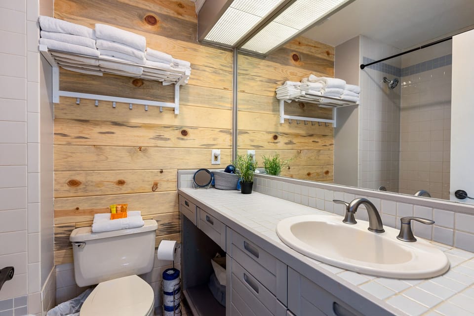 A bathroom with wood-paneled walls, a large mirror, white tiled countertop, sink, toilet, and towel shelves.