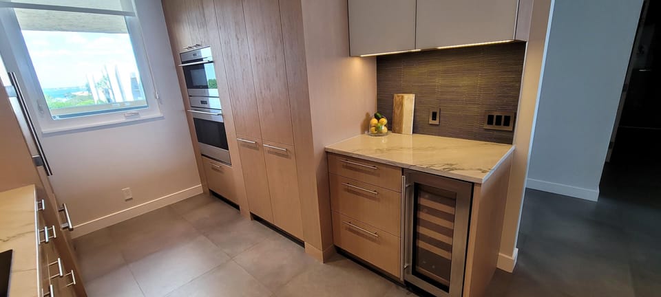 Kitchen corner featuring light wood cabinetry, marble countertop, built-in wine cooler, and textured dark backsplash undercabinet lighting.