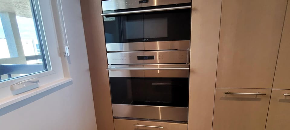 Built-in stainless steel Wolf microwave and oven are set within smooth beige cabinetry beside a window.