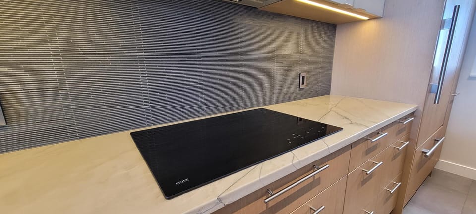 Kitchen countertop with sleek black induction cooktop, beige marble surface, and linear gray tile backsplash.