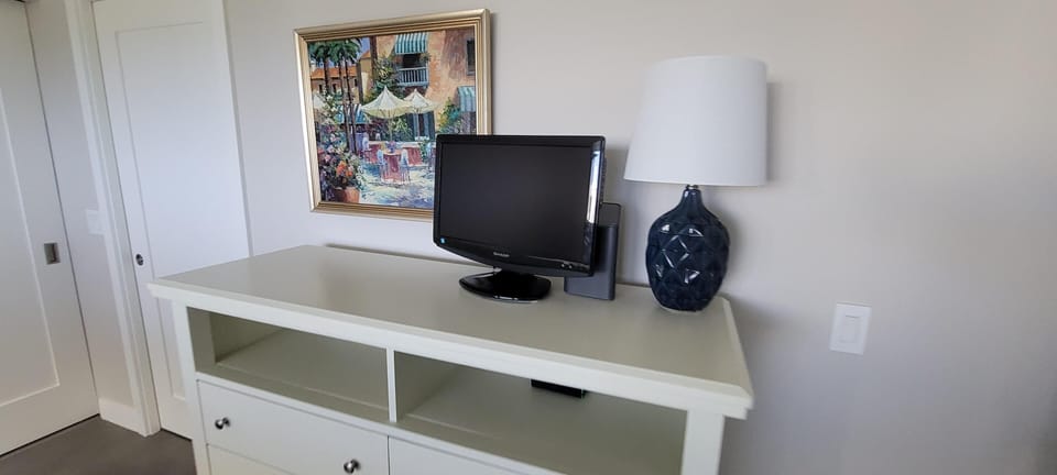 White dresser topped with a small black TV and a dark blue textured ceramic lamp, beneath a colorful framed painting.