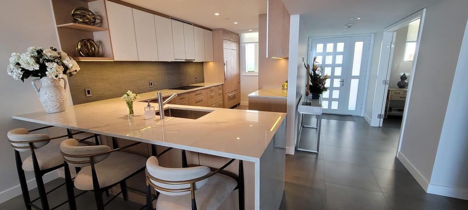 Marble kitchen island with built-in sink and beige cushioned bar stools under recessed lighting.