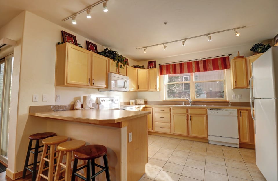 A kitchen with light wood cabinets, a countertop with three wooden stools, white appliances including a refrigerator, stove, and dishwasher, and a window with a red striped valance.
