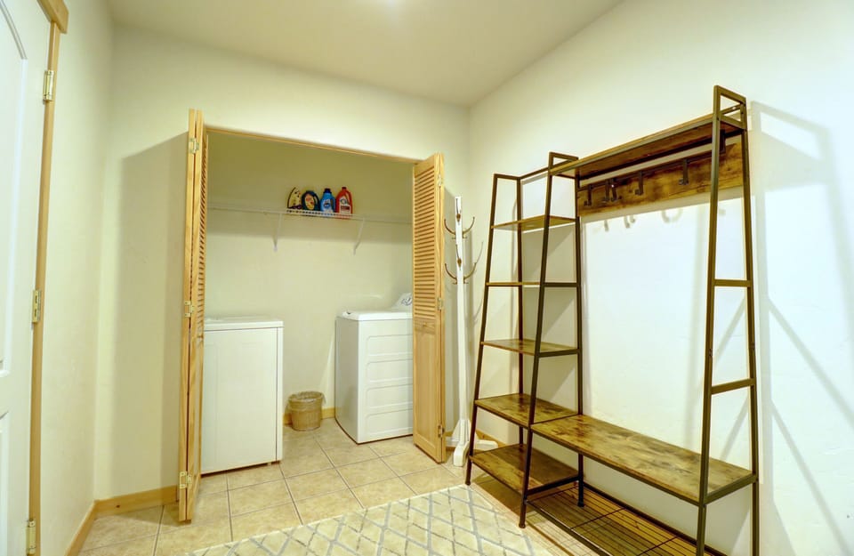 A laundry room with a washer, dryer, shelf with cleaning supplies, a wooden coat rack, and a metal shelving unit. The tiled floor has a patterned rug. The room is well-lit with white walls.