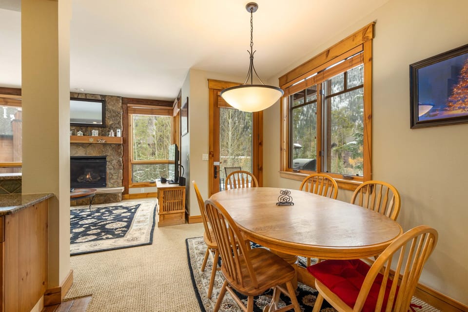 A cozy dining area with a wooden table and six chairs, next to large windows. A fireplace and TV are visible in the adjacent living room.