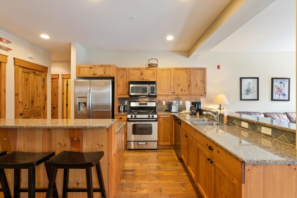 A kitchen features wooden cabinets, stainless steel appliances, a granite countertop with two bar stools, and overhead lighting. Adjacent is a living area with a couch, side table, and framed artworks.
