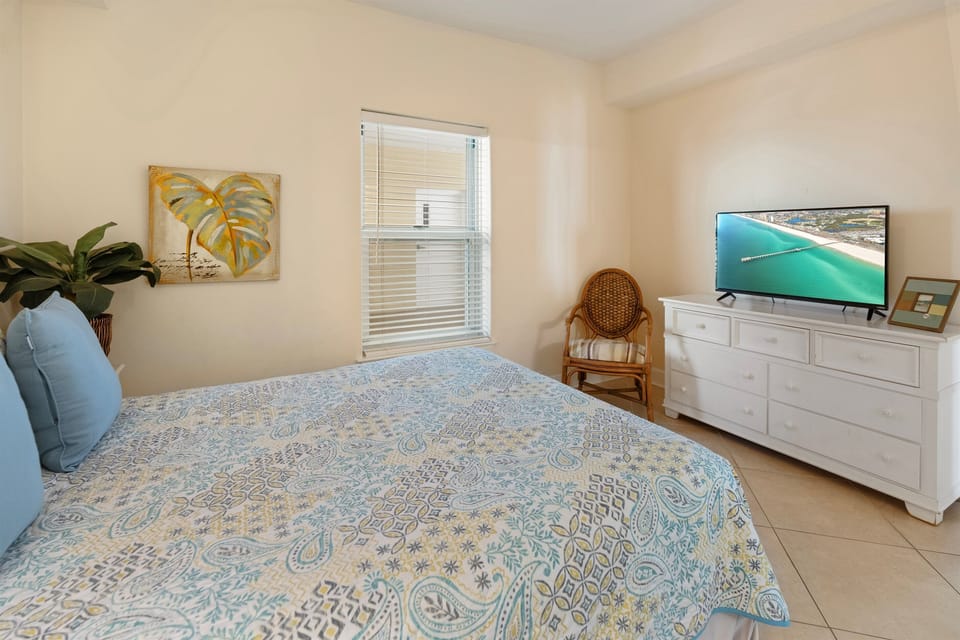 Relax in your bright coastal bedroom with tropical touches, featuring a comfy bed and entertainment setup for peaceful evenings.
