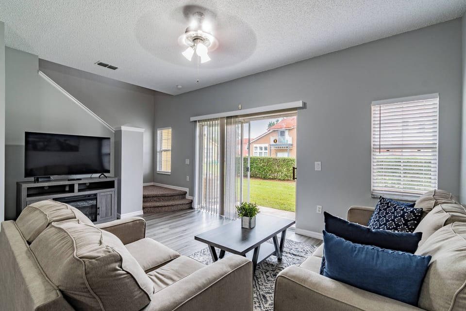 This bright, airy living room boasts cozy beige sofas with deep blue pillows, inviting relaxation. Large sliding doors reveal a lush outdoor view, filling the space with natural light and fresh air for your enjoyment.