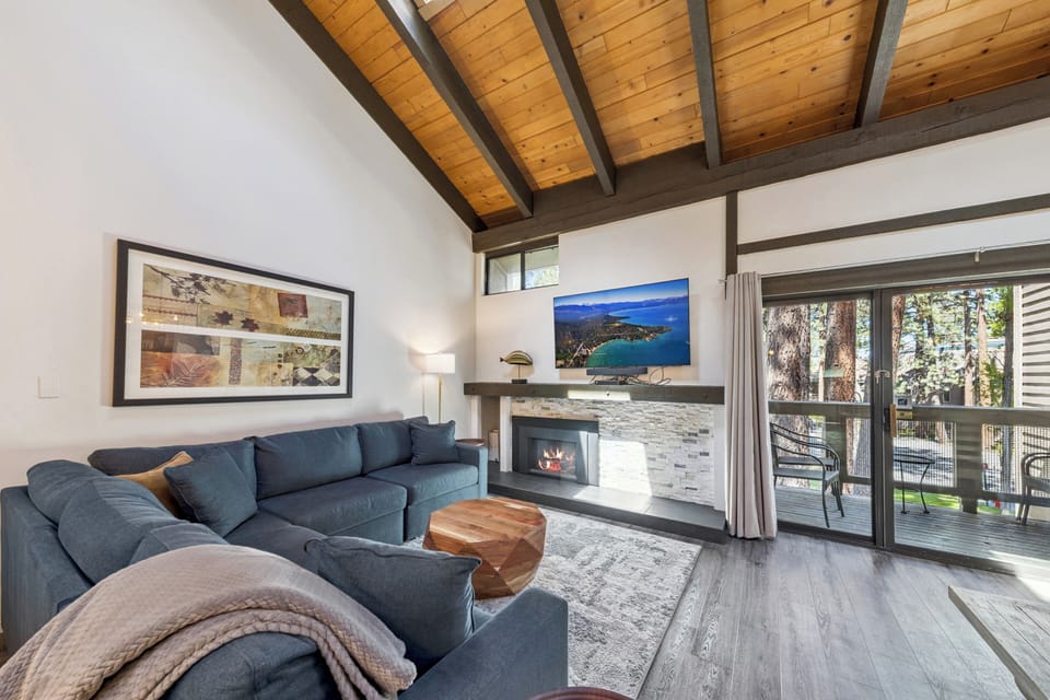 Sink into your plush sectional sofa beneath soaring wood-beamed ceilings and let warmth from the fireplace embrace you. Natural light floods through expansive windows, framing forest views beyond your private deck—your perfect mountain retreat awaits.