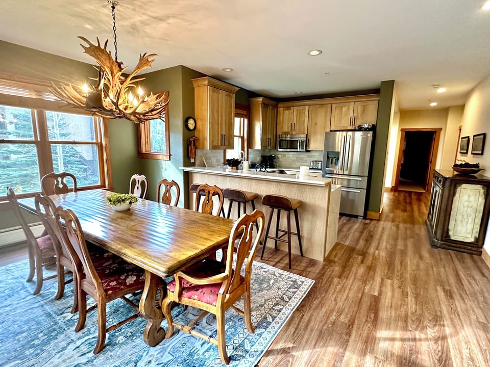 Dining room with a large wooden table and chairs, an antler chandelier, a kitchen with modern appliances, and wooden flooring.