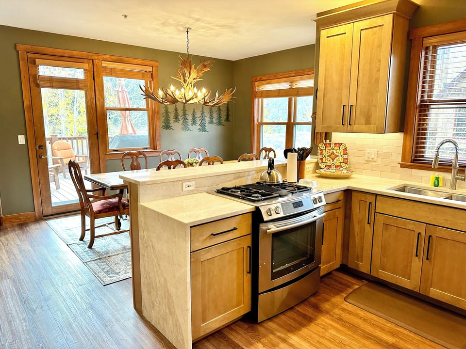 Kitchen and dining area with wooden cabinets, stainless steel stove, and sink. Dining table set near a window. Deer antler chandelier above table. Door to outside visible.