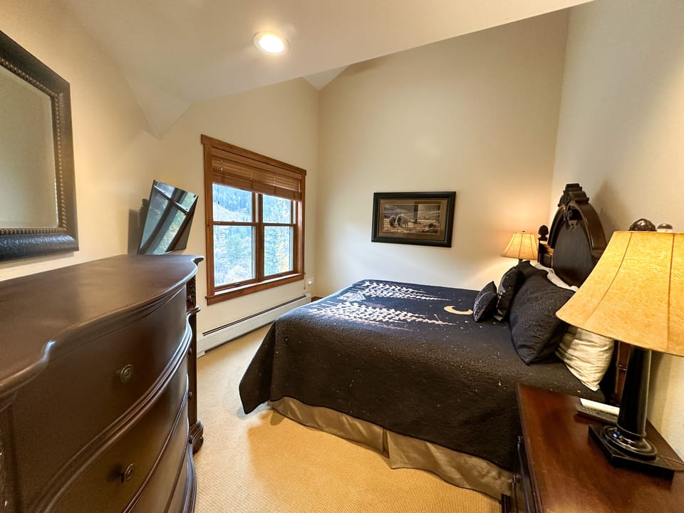 Cozy bedroom with a double bed, two bedside lamps, a landscape painting, a dresser with mirror, and a large window with wooden blinds. The walls are light-colored.