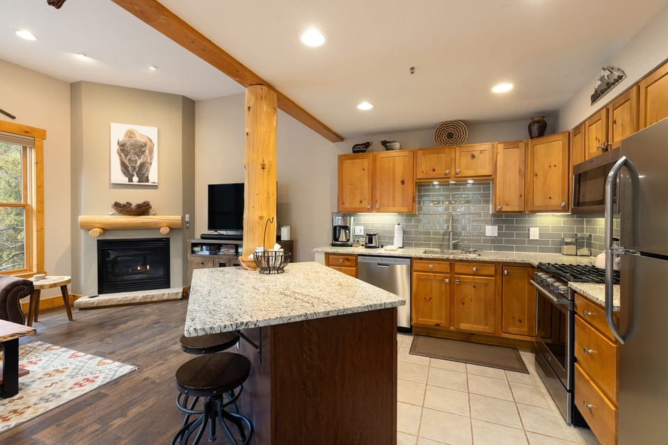 A cozy kitchen with wooden cabinets, granite countertops, and stainless steel appliances. There's a kitchen island with bar stools. A living area with a fireplace and TV is visible in the background.