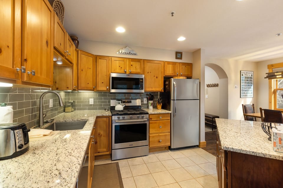 A modern kitchen with wooden cabinets, stainless steel appliances, granite countertops, and a tiled backsplash. There is a microwave, stove, refrigerator, and a toaster on the counter.