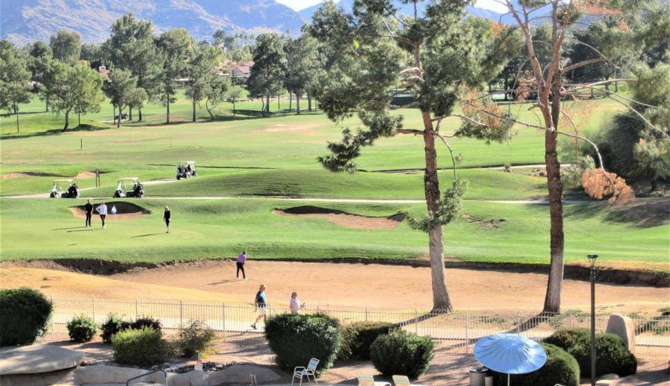 Clear blue pool with curved edges and lounge chairs shaded by a blue umbrella, overlooking green golf course.