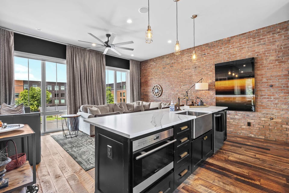 Spacious and inviting living room featuring comfortable sectional seating, exposed brick walls, a Smart TV, and an open-concept layout that flows seamlessly into the kitchen and dining area.
