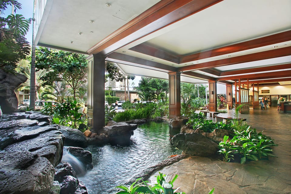 This property features a tranquil covered entrance with a serene koi pond, tropical landscaping, and wooden beam accents connecting the outdoor garden to the indoor reception area.