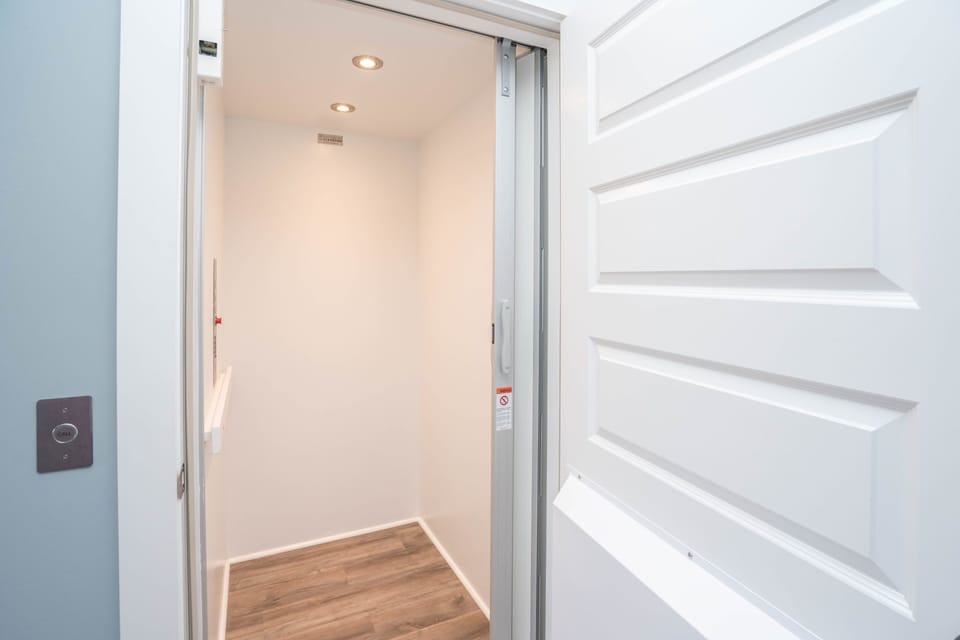 Private residential elevator with open white door, call button, recessed lights, handrail, wood-look flooring, and easy multi-level access.