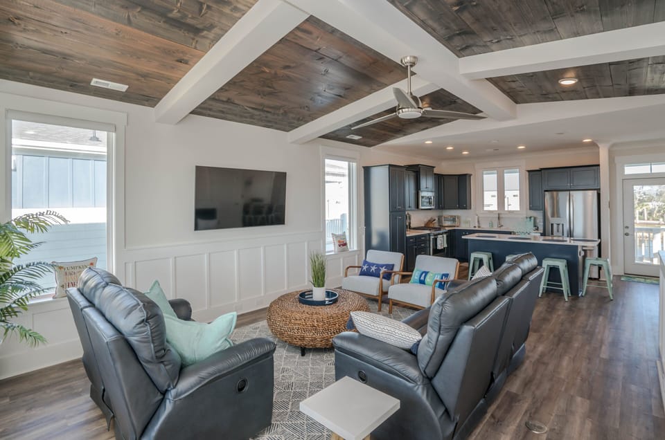 Bright living room with wood-beam ceiling, wall TV, leather seating, accent chairs, round woven coffee table, ceiling fan, large windows, and deck access.