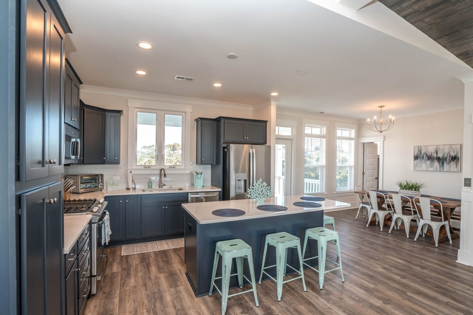Modern kitchen with navy cabinets, large island, mint stools, stainless appliances, gas range, dining table, chandelier, tall windows, and wood floors.