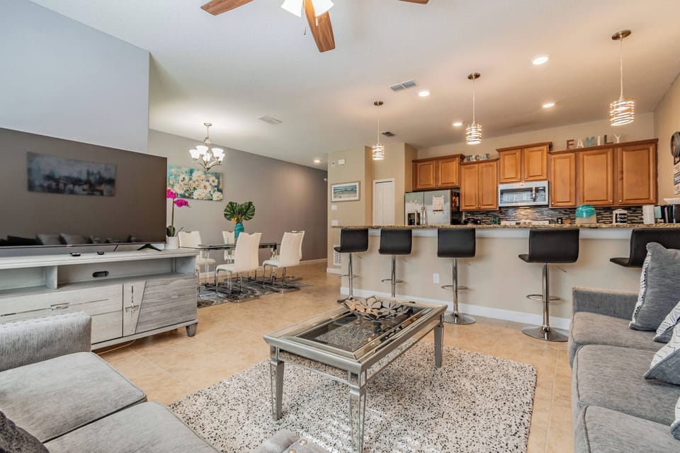 Enjoy a bright and spacious living area filled with natural light, featuring a stylish grey sectional and elegant glass coffee table, all seamlessly connected to a modern kitchen perfect for relaxing and socializing with loved.